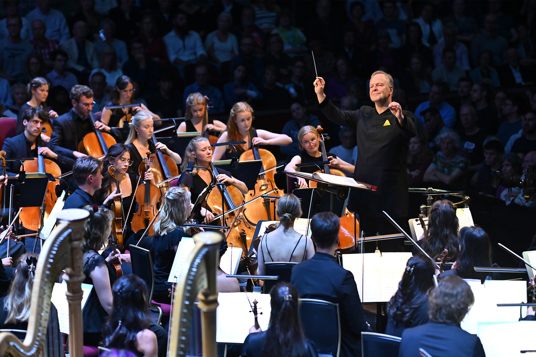 A man wearing a conductor's suit, conducting an orchestra with his hands, with an orchestra performing on stage, with an audience behind, all wearing smart attire.